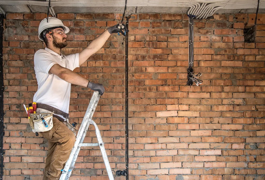Operari electricista amb casc i cinturó d'eines instal·lant el cablejat elèctric sobre una paret de maó vist durant una reforma integral. - Equip Cairat Reformes